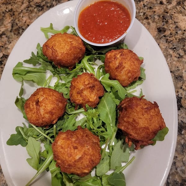 Six golden-brown fried arancini balls on a bed of arugula, with a side of red marinara sauce.