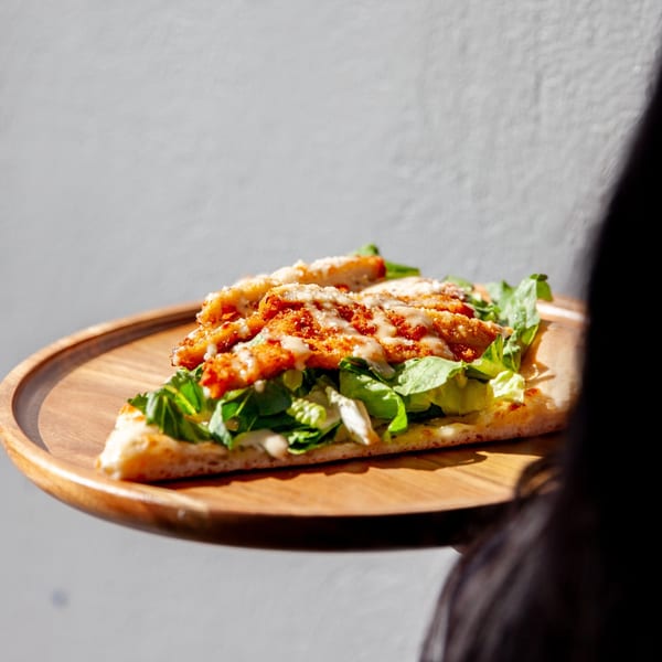 A slice of Sayville Pizza with crispy chicken, fresh lettuce, and creamy dressing on a wooden plate.